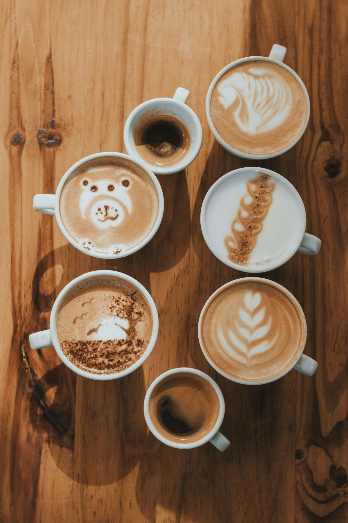 Several cups of cappuccino with latte art and two espressos seen from above on a wooden table